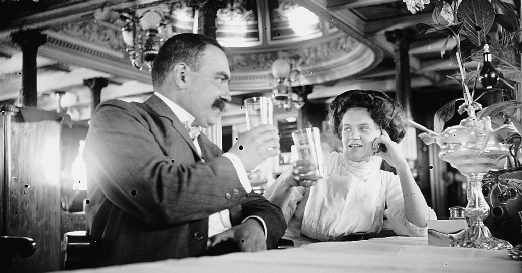 Svartvitt foto av en man och en kvinna som skålar med varandra i ett elegant inrett rum med ornamenterade tak och taklampor. Mannen, med mustasch, bär kostym, medan kvinnan har en vit blus och uppsatt hår.