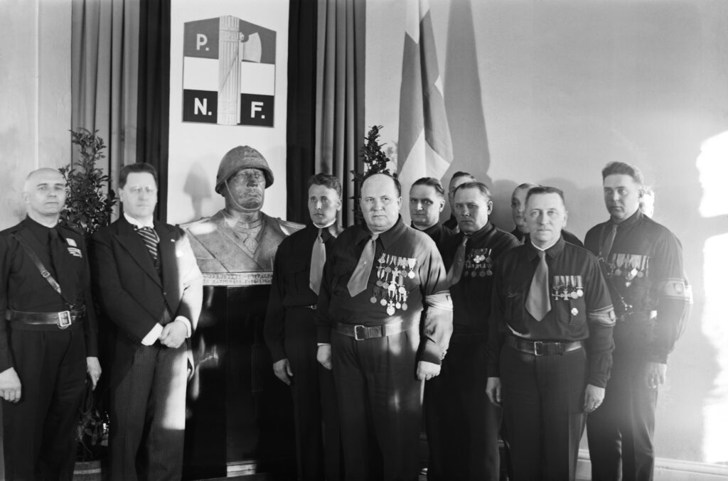 Svartvitt foto av en grupp män i militärliknande uniformer står framför en byst.