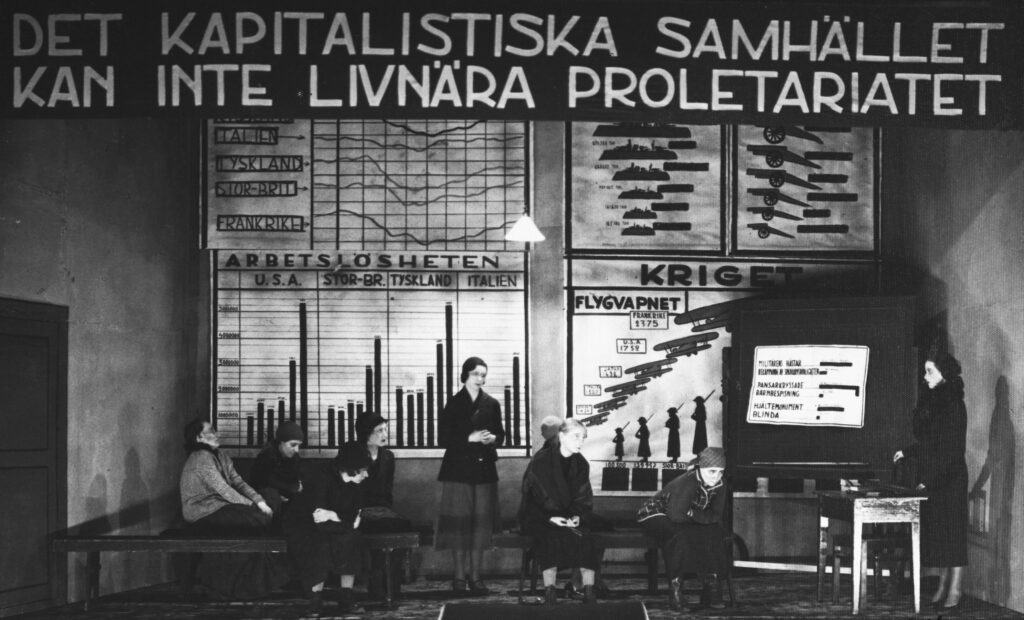 Svartvitt foto av en teaterscen där åtta kvinnor är på scenen. Två av kvinnorna står medan resten sitter.