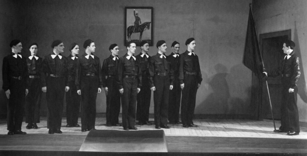 Svartvitt foto på en grupp män klädda i uniformsliknande dräkter och baskrar på huvudet. En av männen håller i en svart flagga. I bakgrunden syns en tavla med en man till häst med höjd arm.