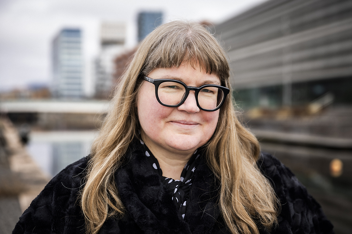 Färgfoto av en kvinna med långt ljust hår och stora svarta glasögon, klädd i svart, stående utomhus framför moderna byggnader och en vattenspegel.