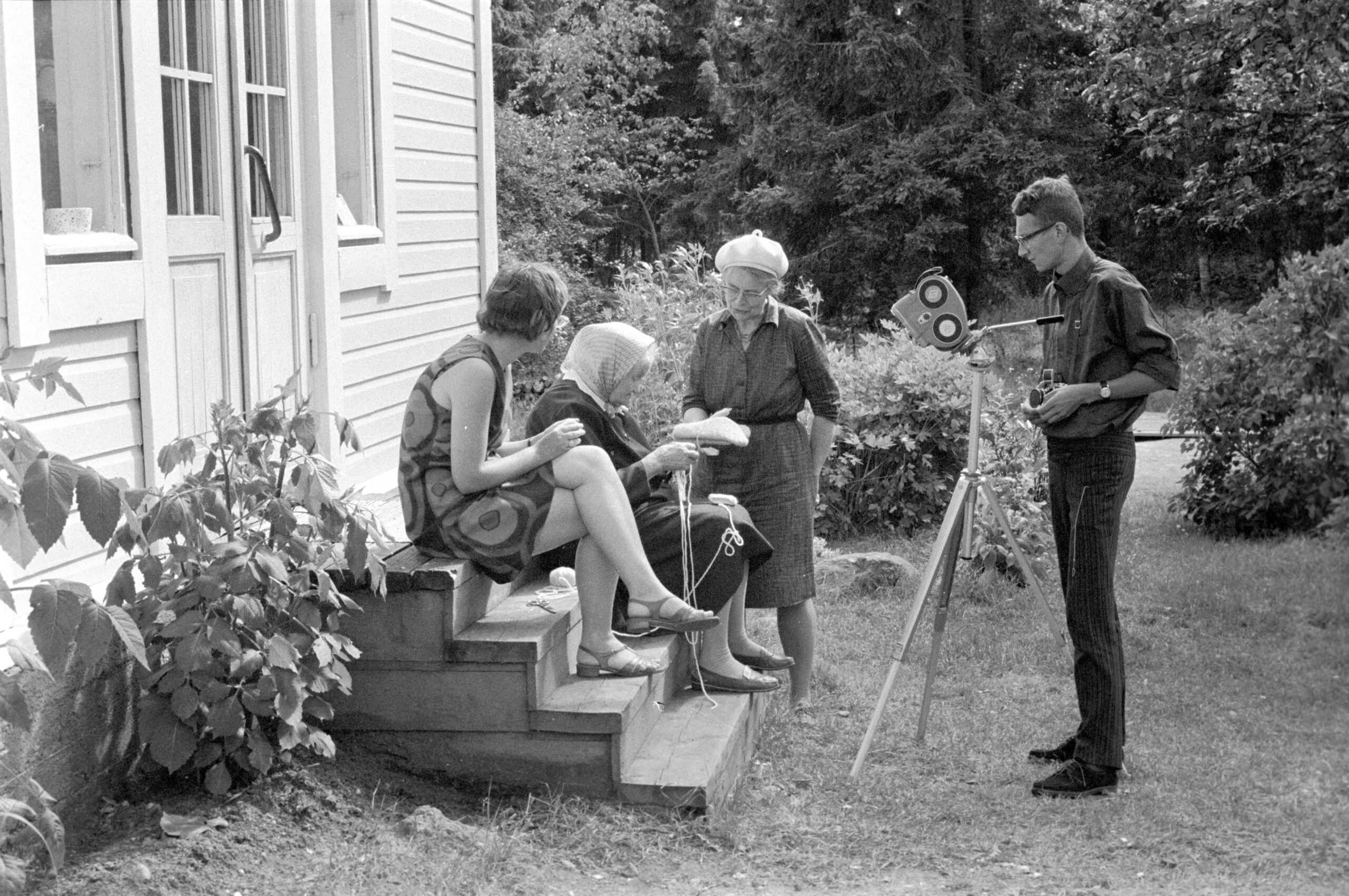 En man filmar tre handarbetande kvinnor som sitter på en hustrappa