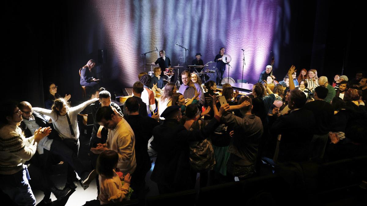 Foto av ett scenframträdande där ett band spelar live medan en stor grupp glada människor dansar, skrattar och applåderar framför scenen i färgrikt scenljus.