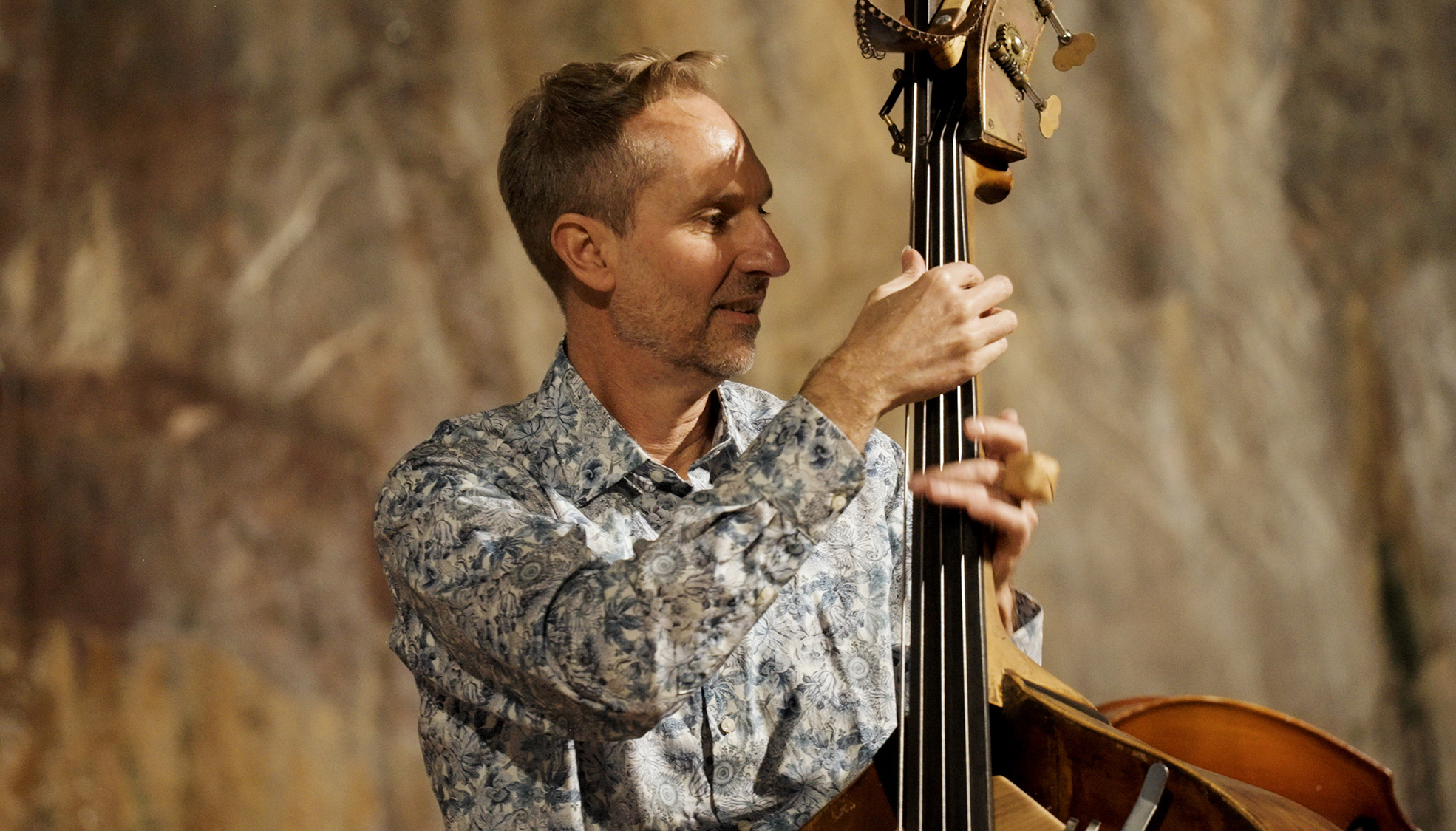 Värivalokuva hymyilevästä miehestä, jolla on kuvioitu paita ja joka soittaa kontrabassoa epätarkkaa, lämpimän sävyistä taustaa vasten.