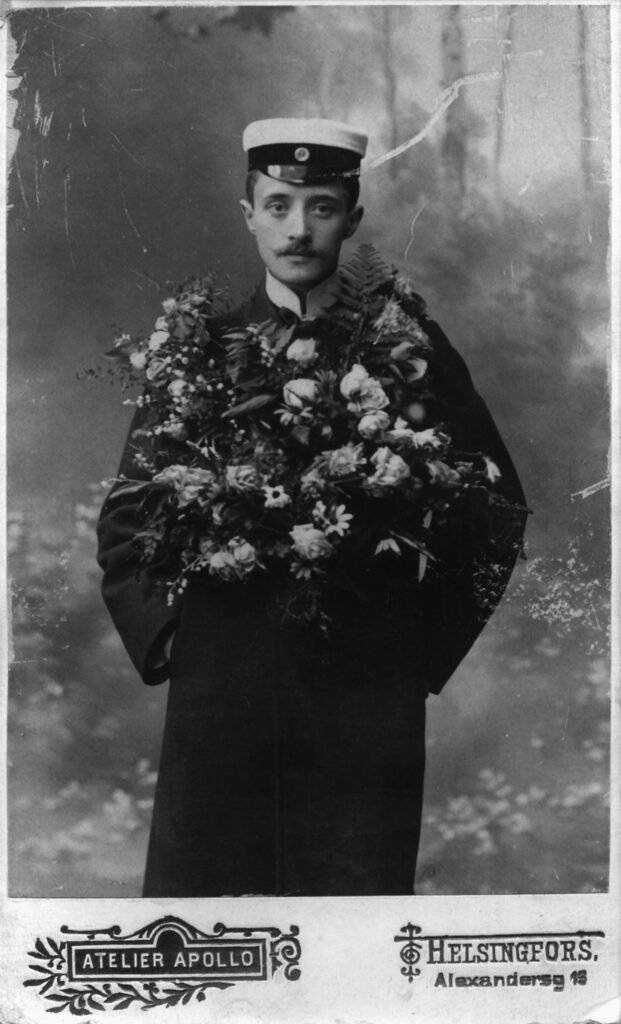 Svartvitt foto av en ung man i studentmössa och mörk rock, som poserar framför neutral bakgrund med en stor blomstergirlang runt axlarna.
