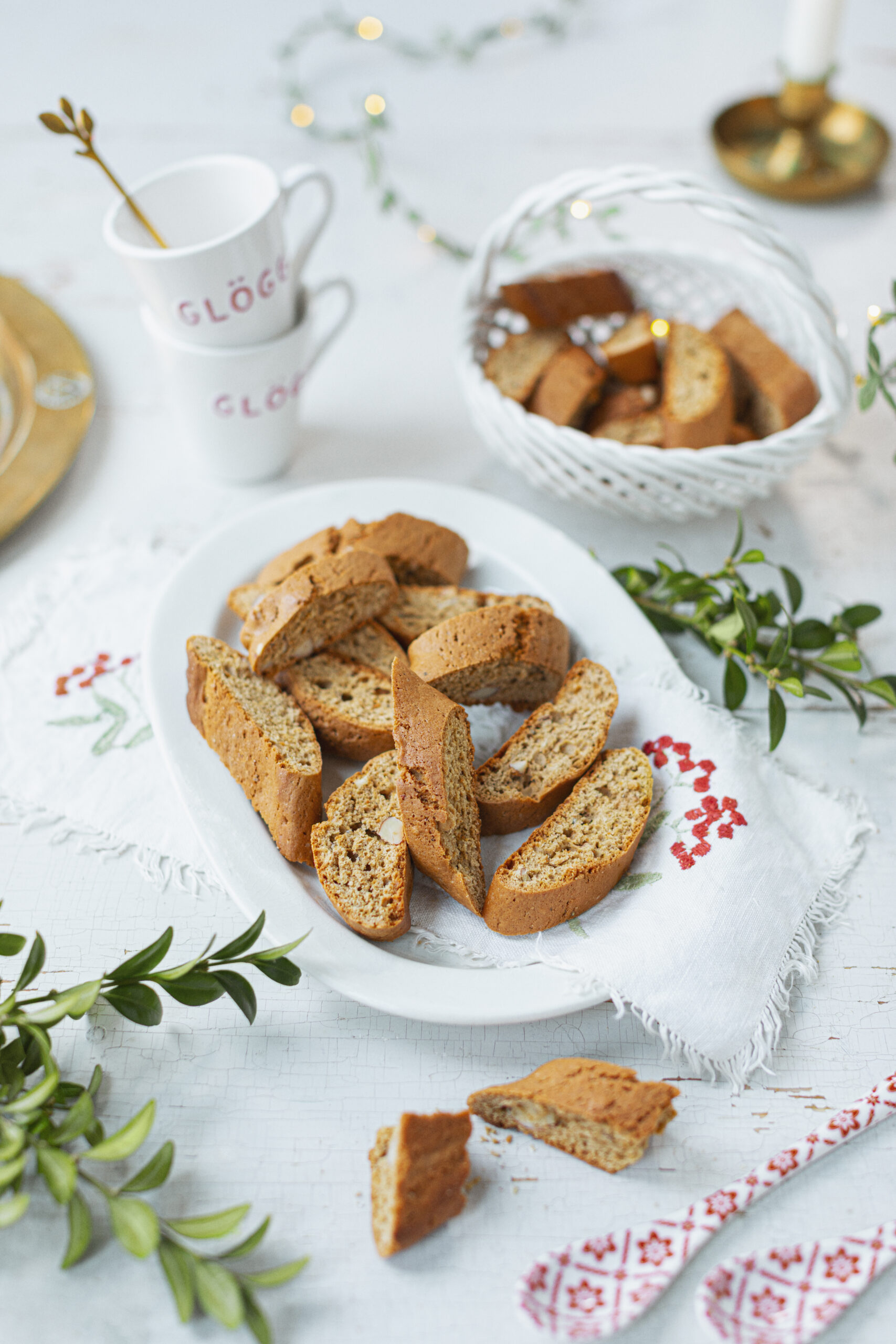 Foto av skivade pepparkaksskorpor på ett vitt fat med broderad duk, gröna kvistar, två staplade glöggmuggar och korg med fler pepparkaksskorpor i bakgrunden.