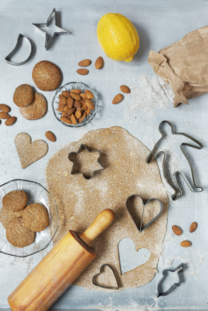 Foto av utkavlad pepparkaksdeg med utstansade former, kakformar, kavel, hela mandlar, citronskal, mjöl och färdiga pepparkakor på en ljus bakgrund.