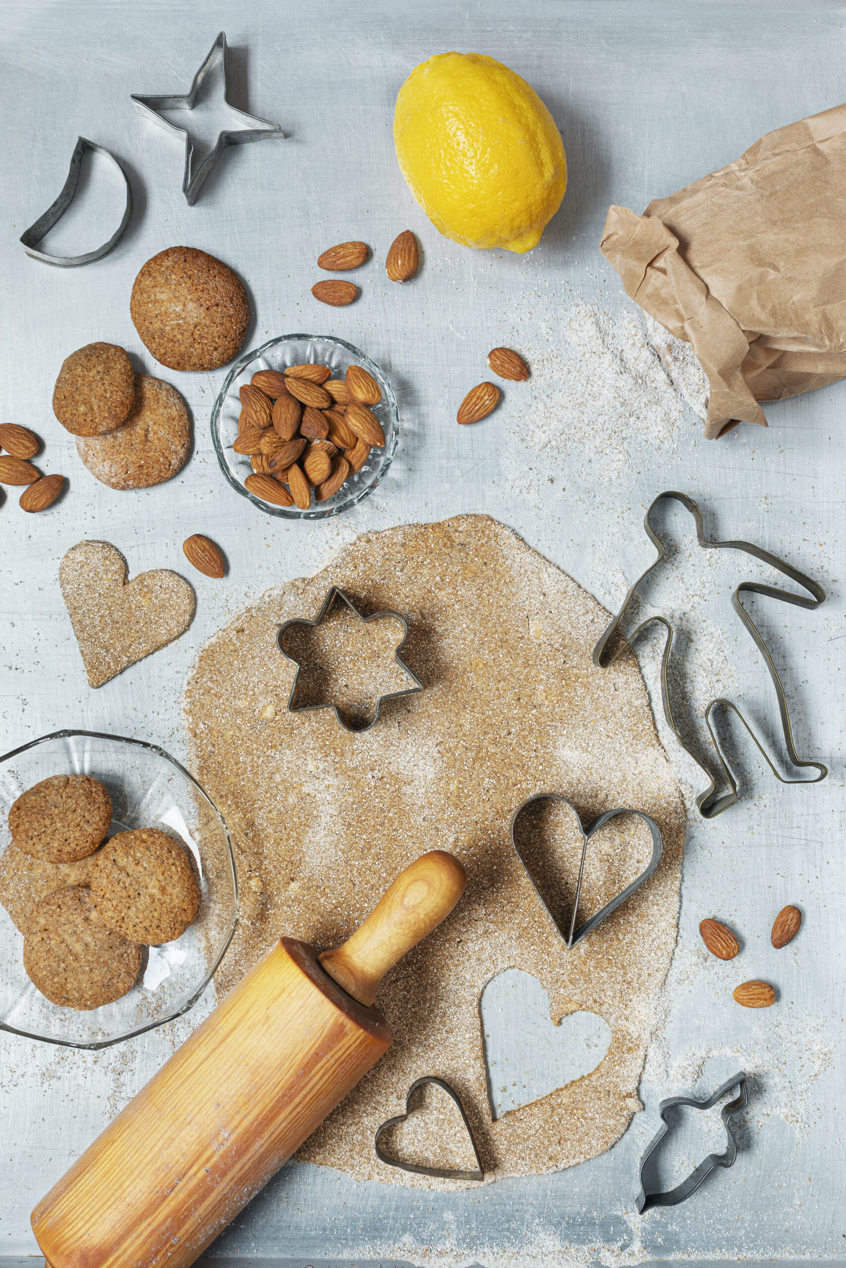 Foto av utkavlad pepparkaksdeg med utstansade former, kakformar, kavel, hela mandlar, citronskal, mjöl och färdiga pepparkakor på en ljus bakgrund.