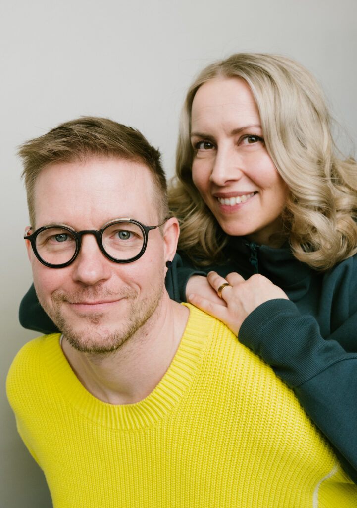 Foto av två leende personer mot ljus bakgrund; mannen har runda glasögon och gul tröja, kvinnan bakom har blont lockigt hår och håller handen på hans axel.